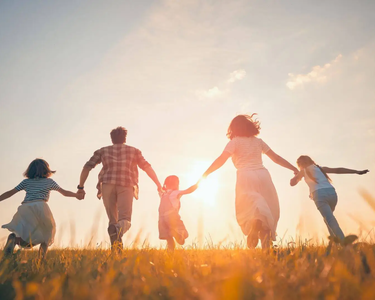Familie mit drei Kindern hält sich an den Händen und läuft gemeinsam über eine Wiese im warmen Licht des Sonnenuntergangs.