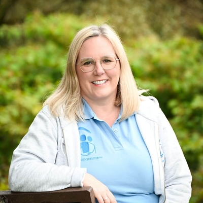 Lächelnde Frau mit Brille sitzt auf einer Holzbank im Grünen und blickt in die Kamera.