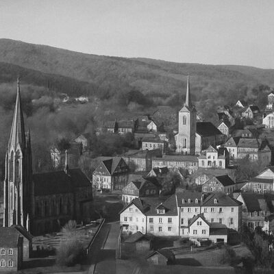 Schwarz-weiß-Fotografie des historischen Stadtkerns der Stadt Kirchen mit zwei Kirchen und traditionellen Fachwerkhäusern, eingebettet in eine hügelige Waldlandschaft.